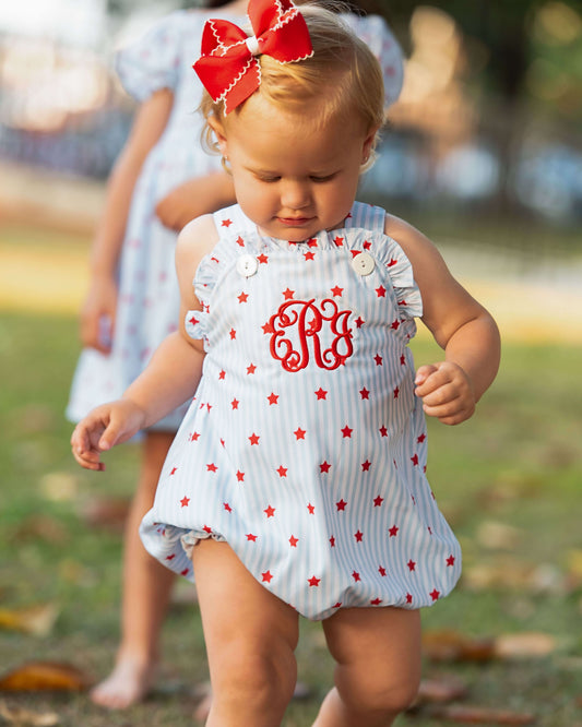 Small child wearing a star print outfit with a monogram on the chest and a red bow on her hair.