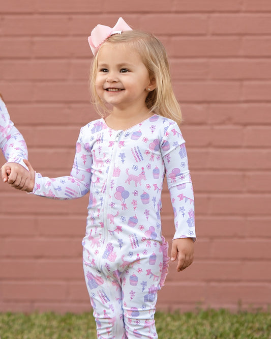 Smiling toddler girl wearing pink and purple birthday print zipper pajamas with balloons, cupcakes, and unicorns, holding hands and standing outdoors against a pink brick wall.