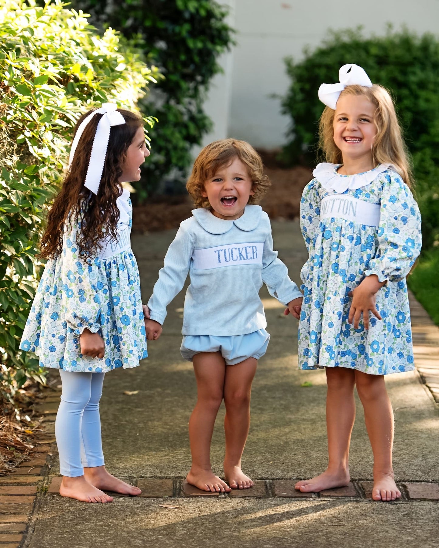 Three children in matching blue floral dresses and light blue pants standing outdoors.