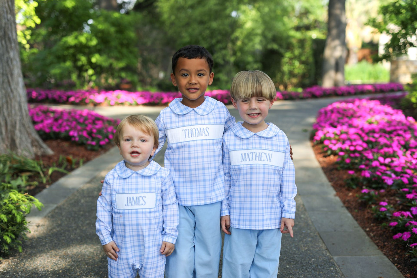 Three children in matching outfits standing on a path with pink flowers and greenery in the background