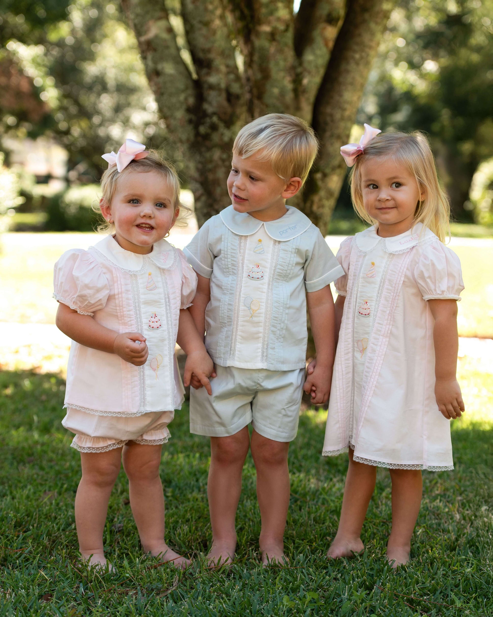 Three children in matching outfits standing on grass with trees in the background