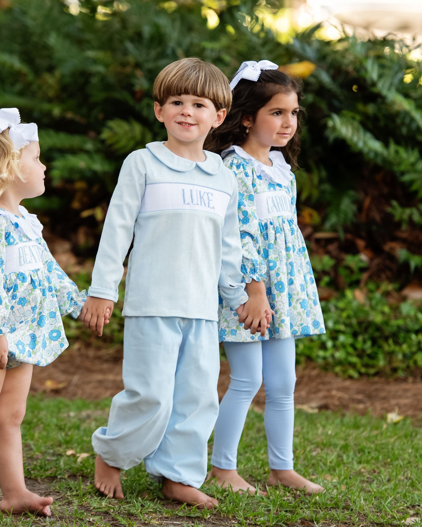 Three children in matching outfits standing outdoors with greenery in the background