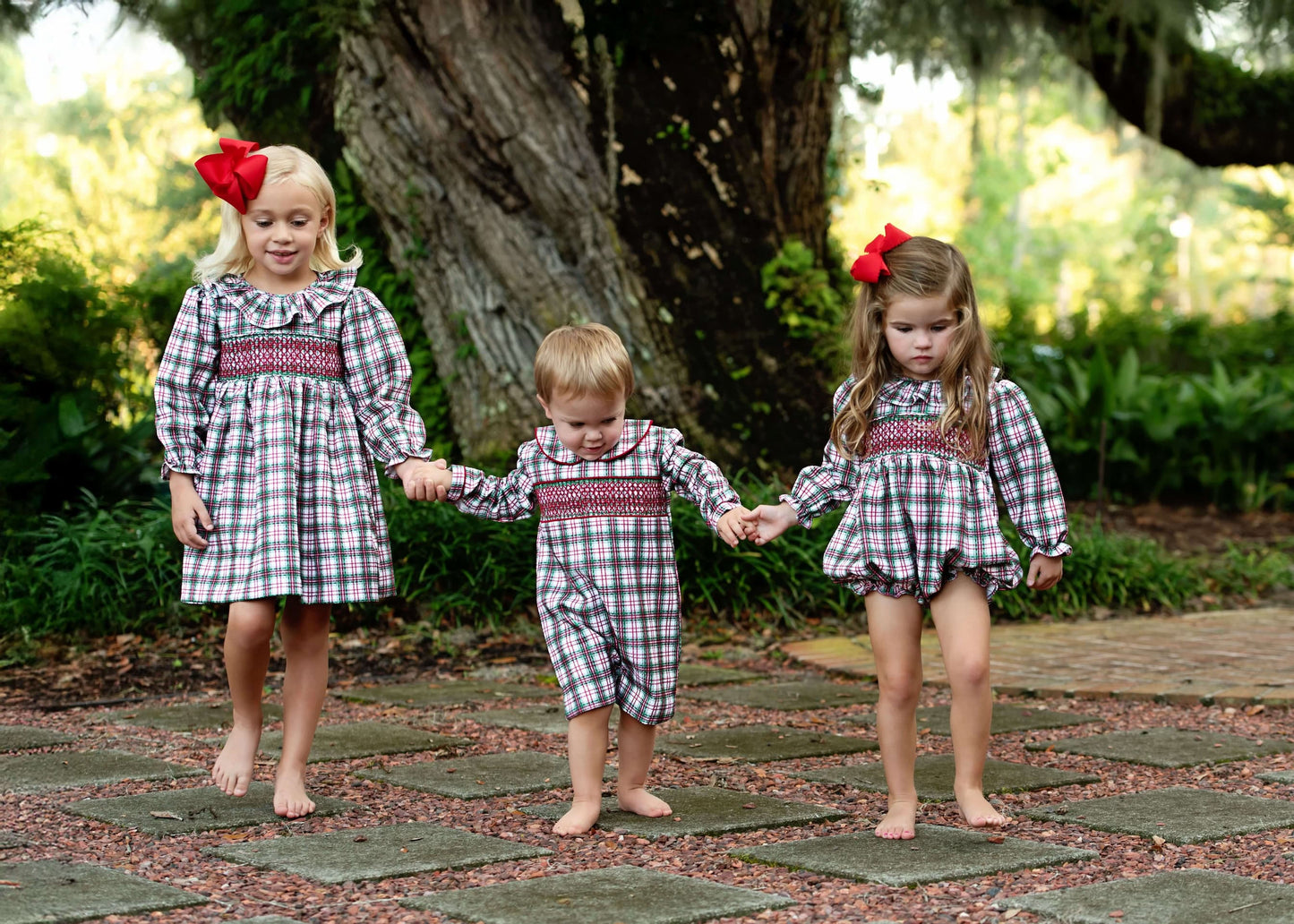 Three children in matching plaid outfits walking hand-in-hand on a stone path.