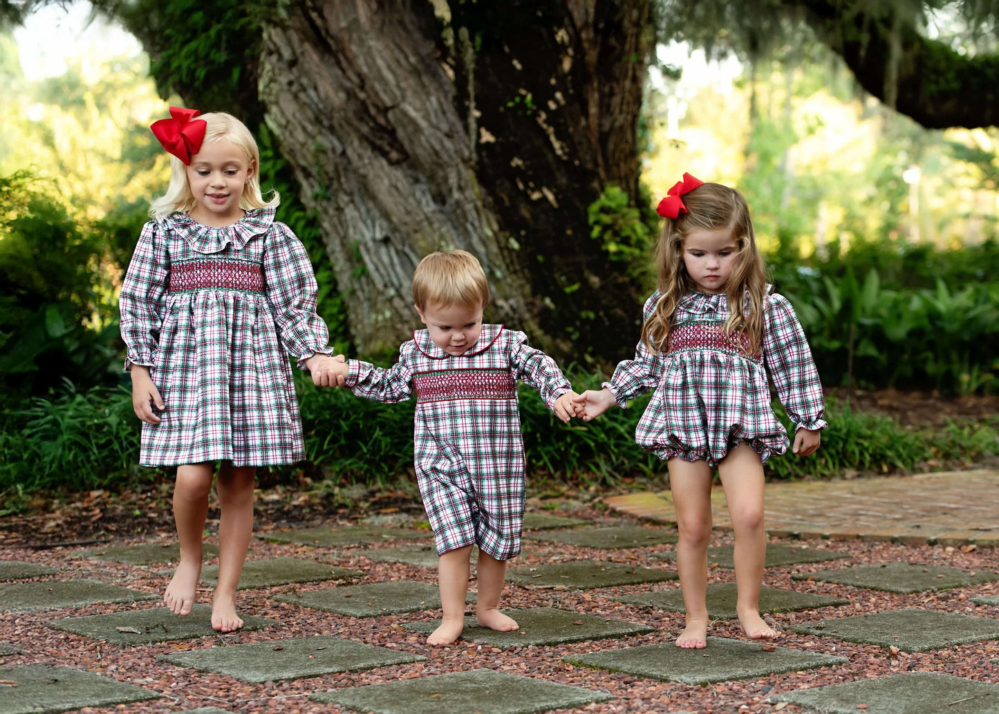 Three children in matching plaid outfits walking hand-in-hand on a stone path.