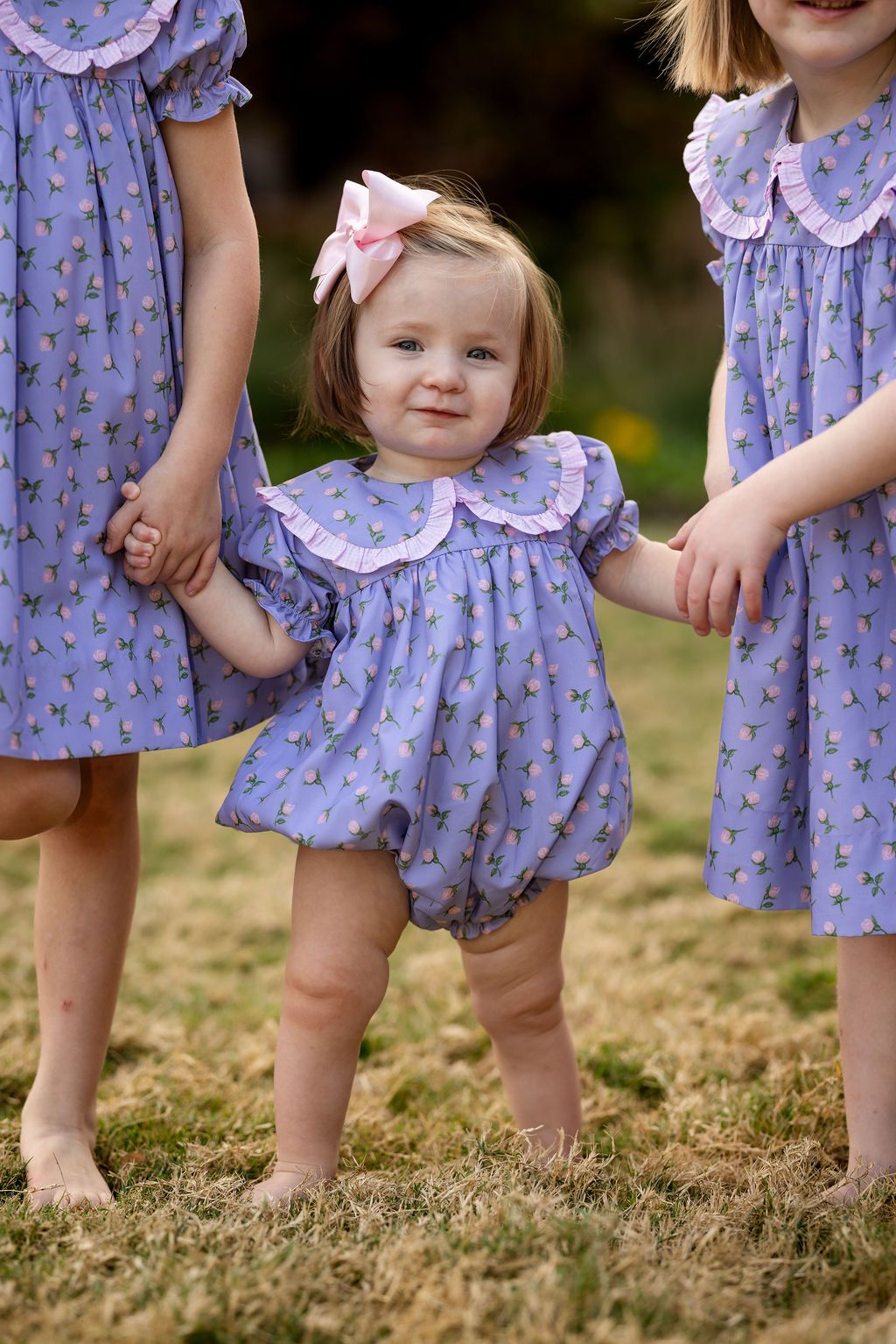 Three children in matching purple dresses standing outdoors on grass.