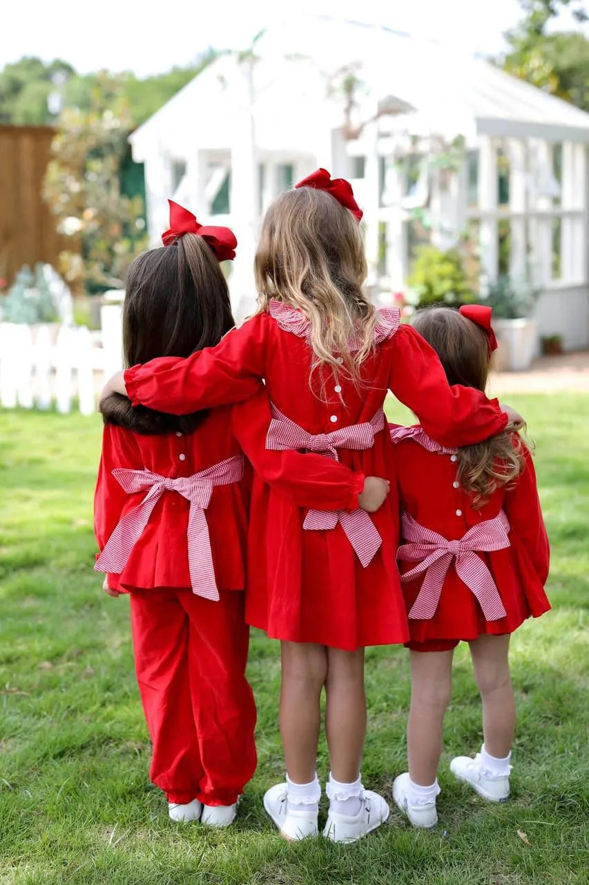Three children in matching red outfits with pink bows standing together outdoors.