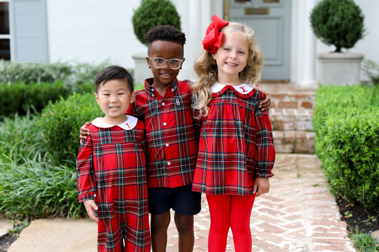 Three children in matching red plaid outfits standing in front of a house.