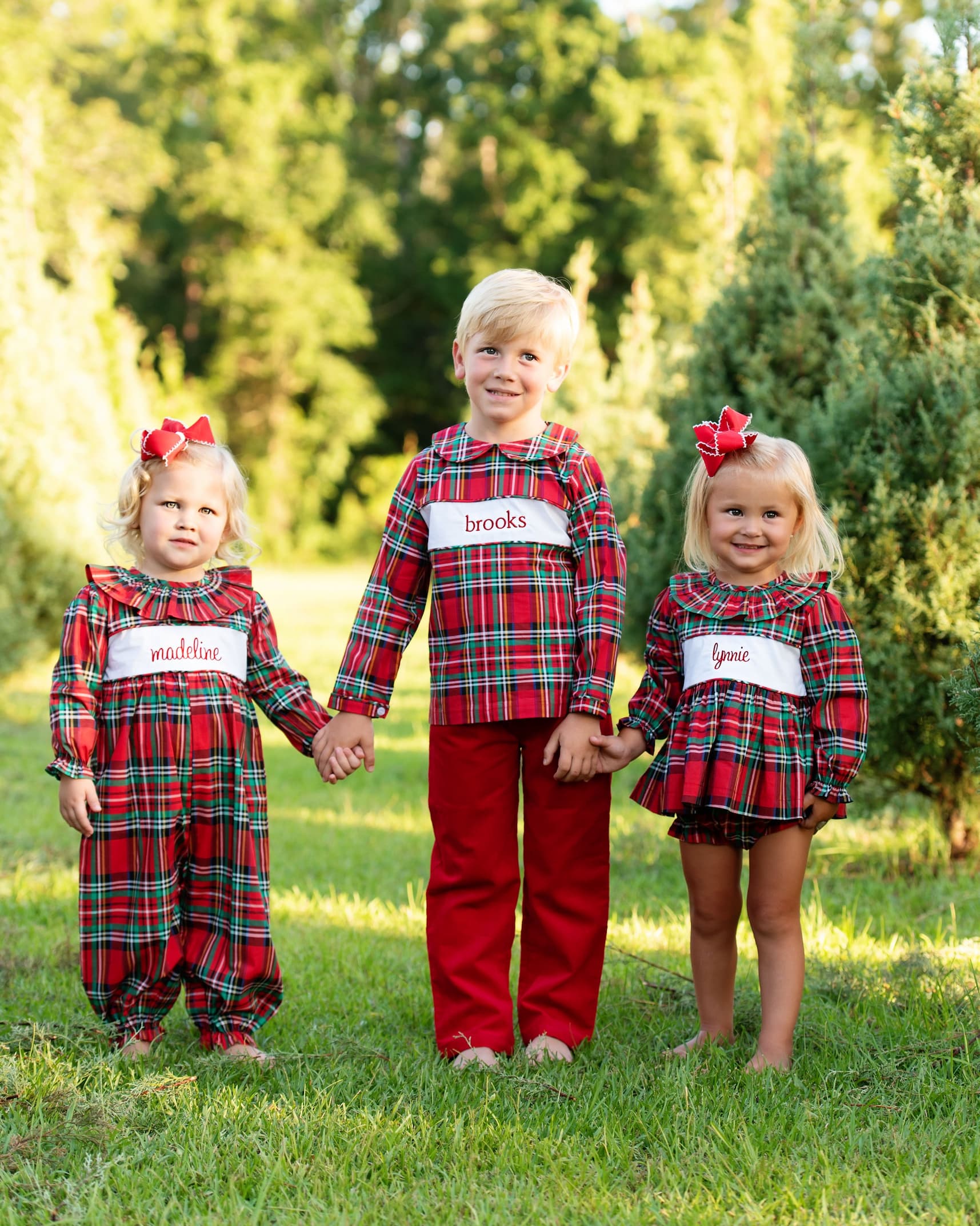 Three children in matching red plaid outfits standing outdoors with trees in the background.