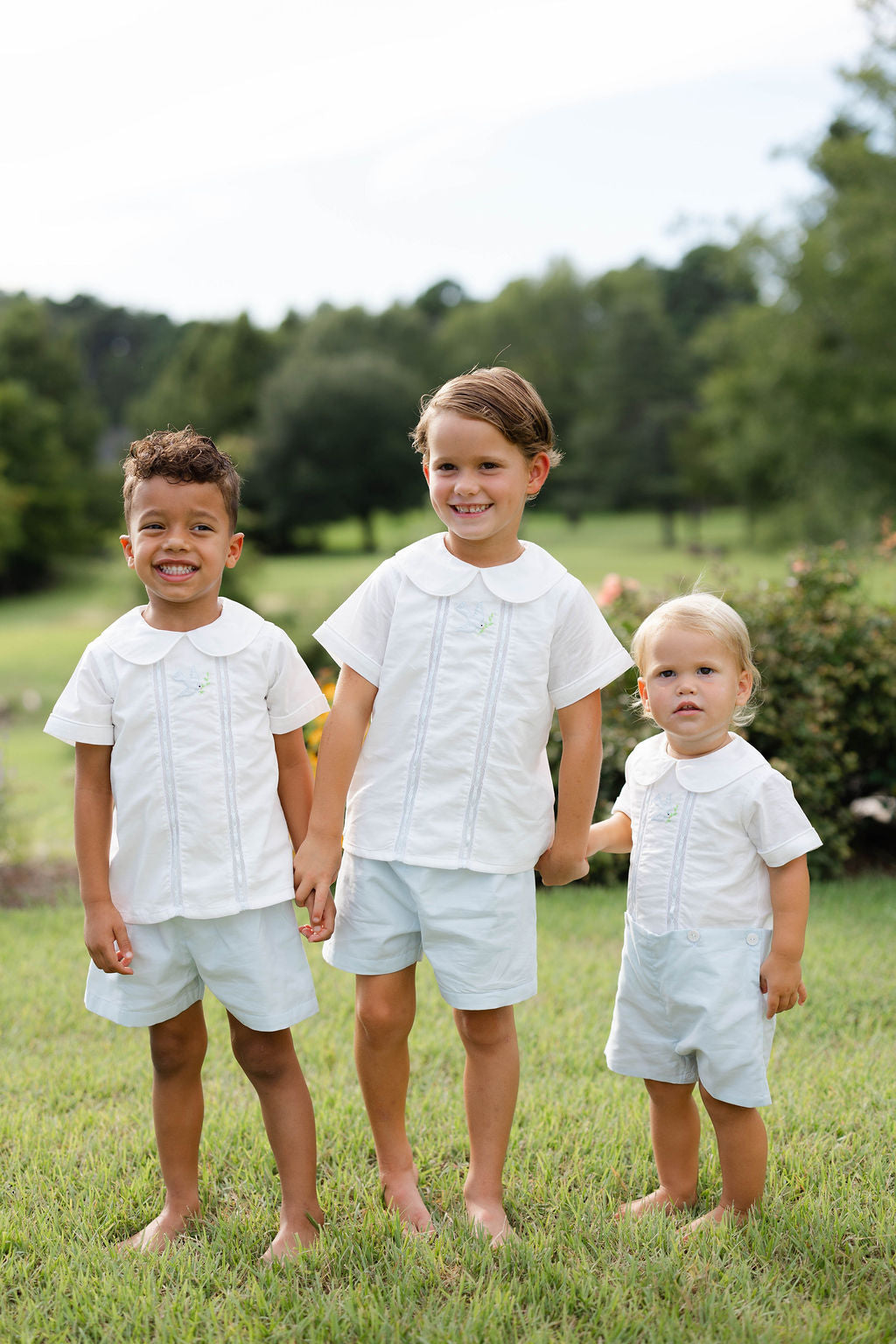Three children in matching white outfits standing in a grassy field with trees in the background.