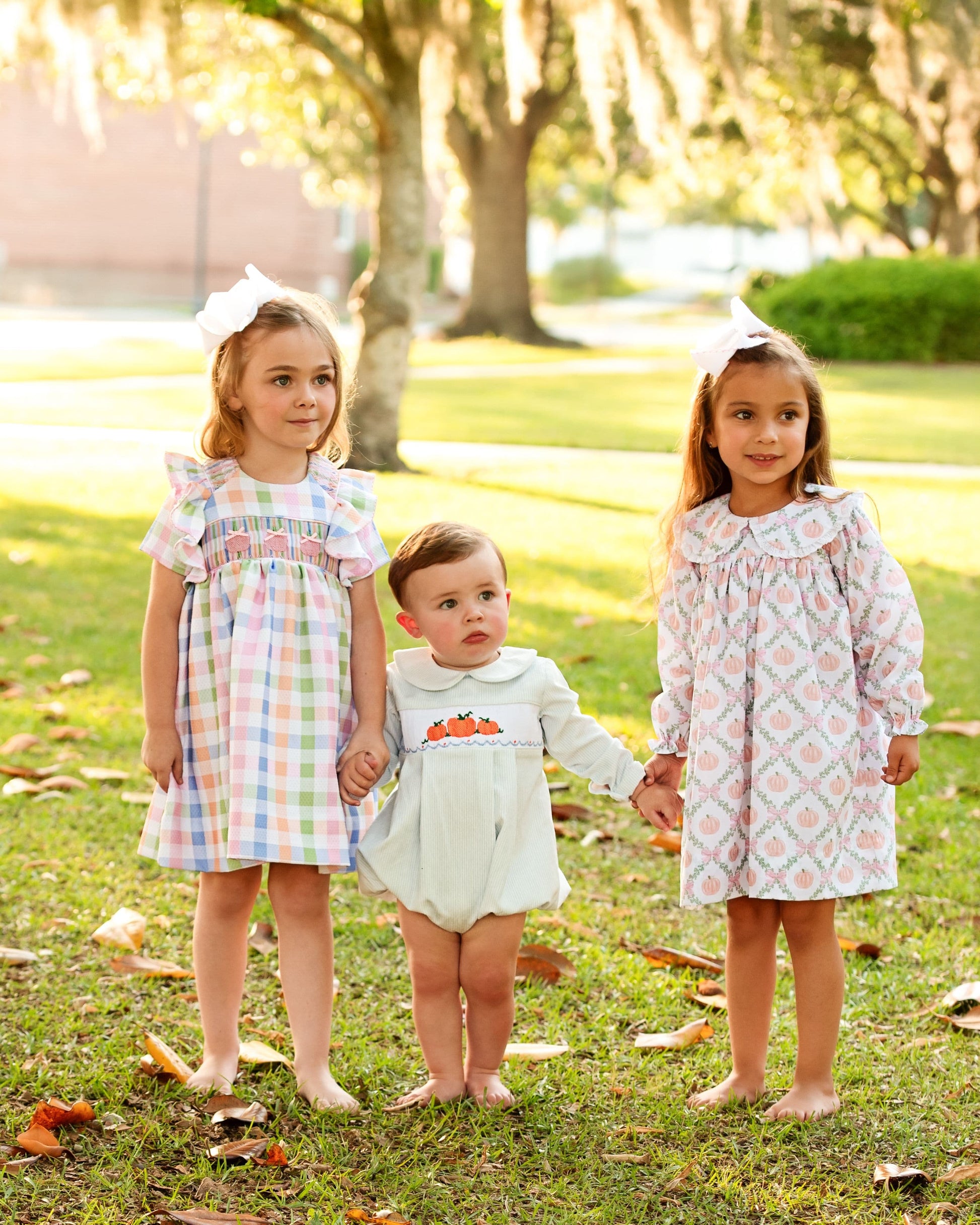Three children in patterned dresses standing on grass with trees in the background
