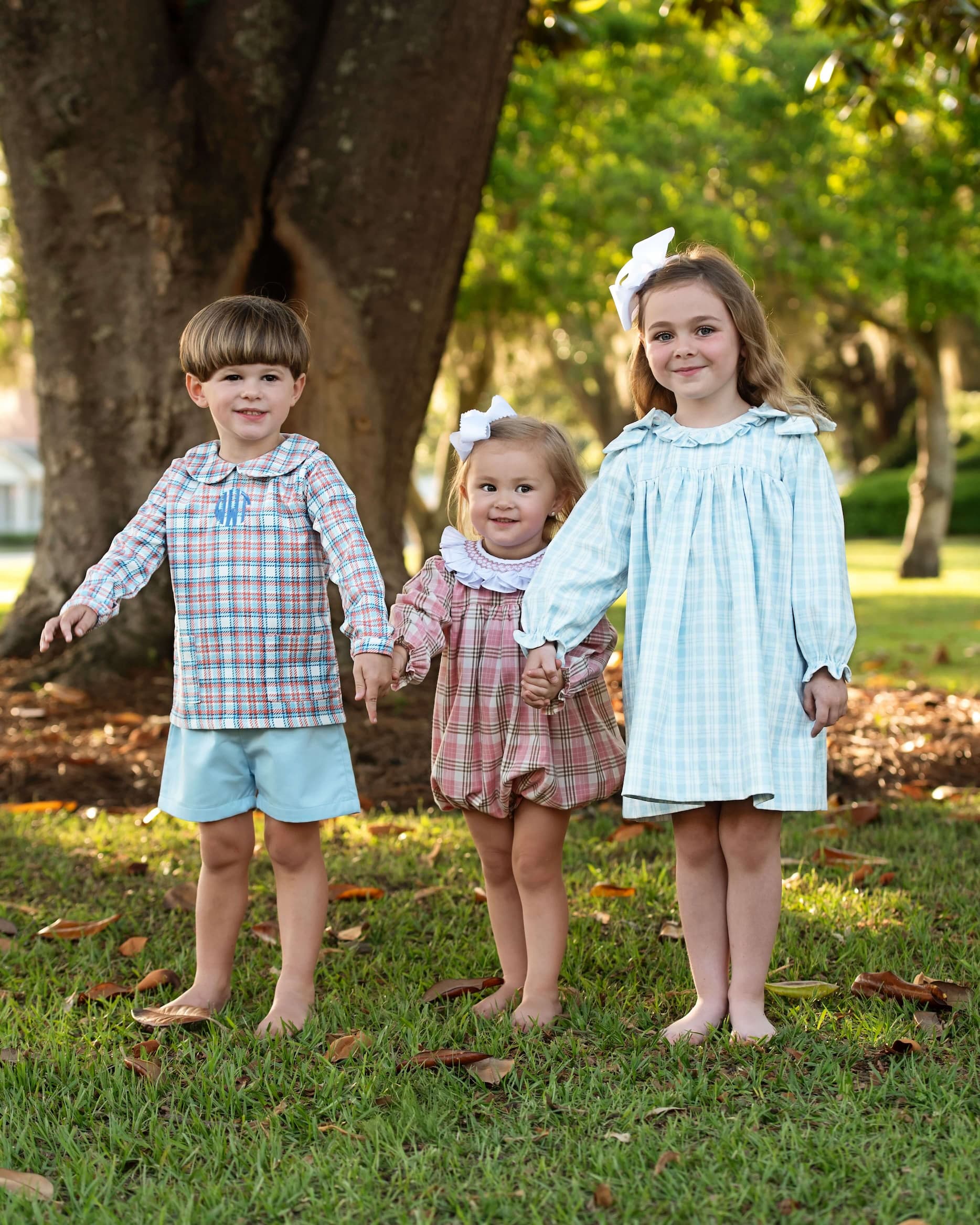 Three children standing outdoors in a park with trees and grass.