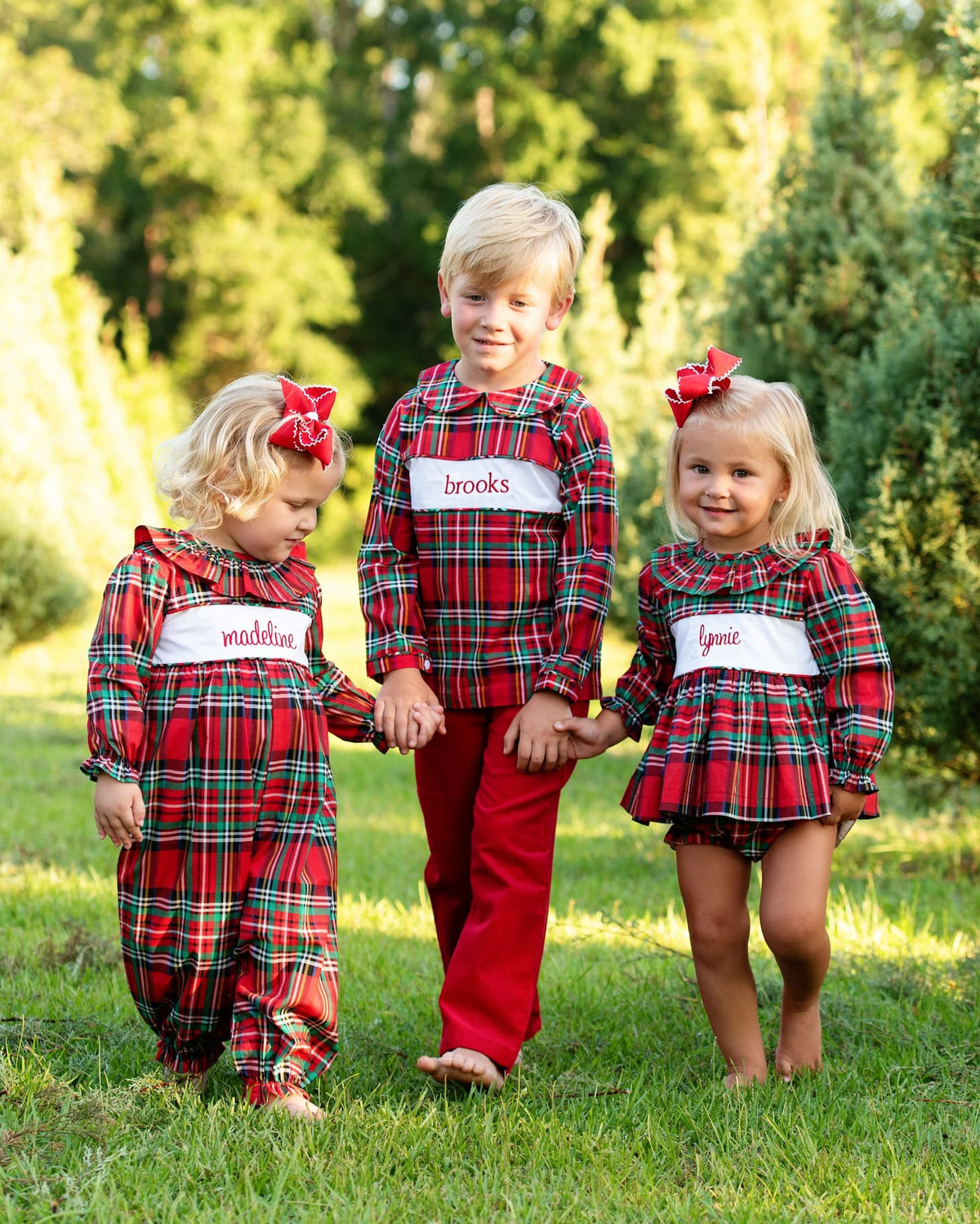 Three children walking hand-in-hand on a grassy lawn wearing matching red and green plaid holiday outfits with white name patches.