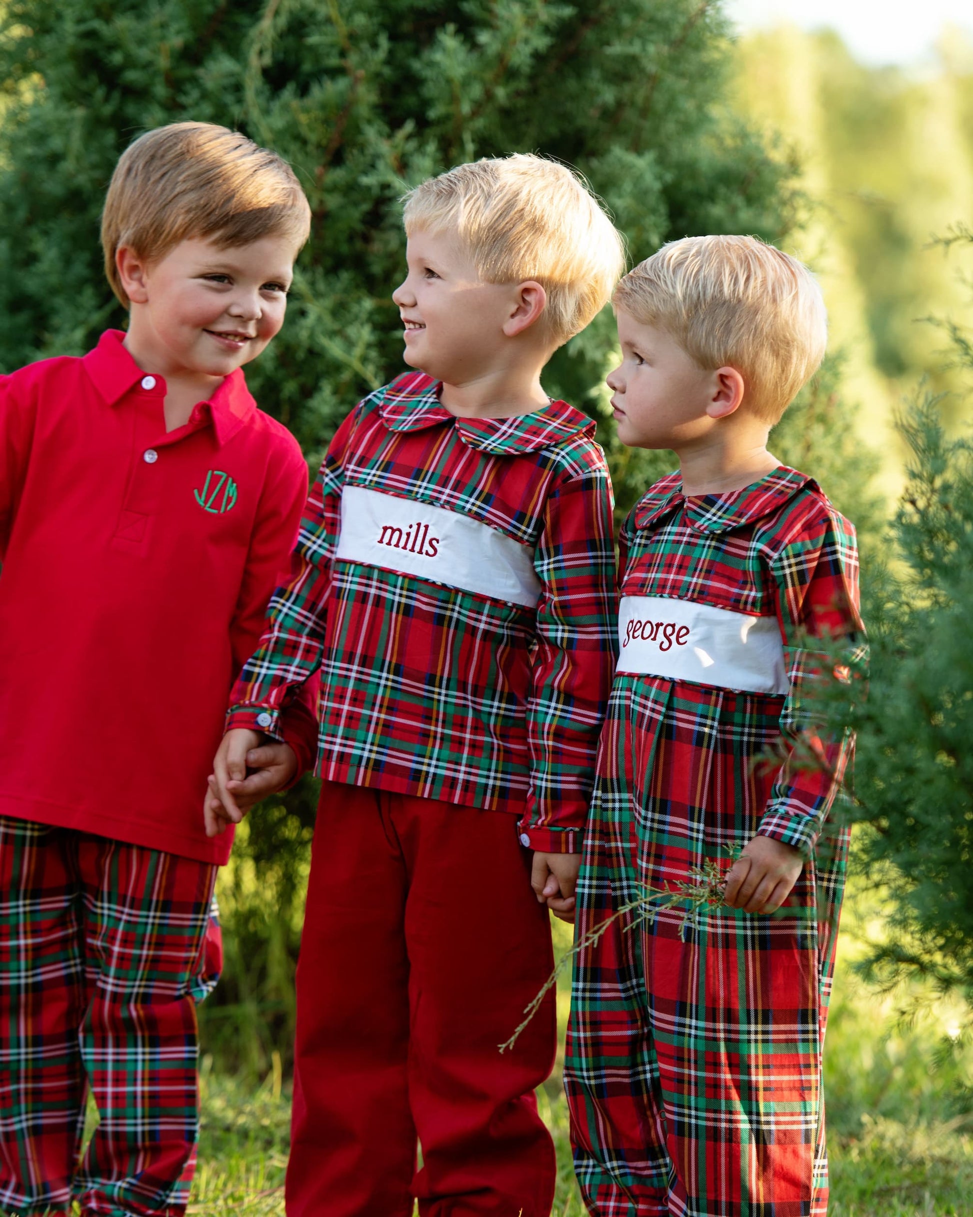 Three children wearing red plaid pajamas with embroidered names in a forest setting.
