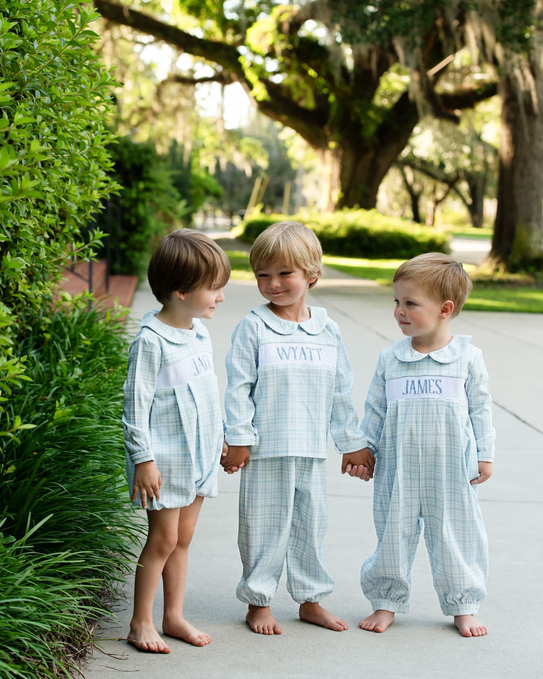 Three young boys in matching outfits standing outdoors on a path with trees and greenery.