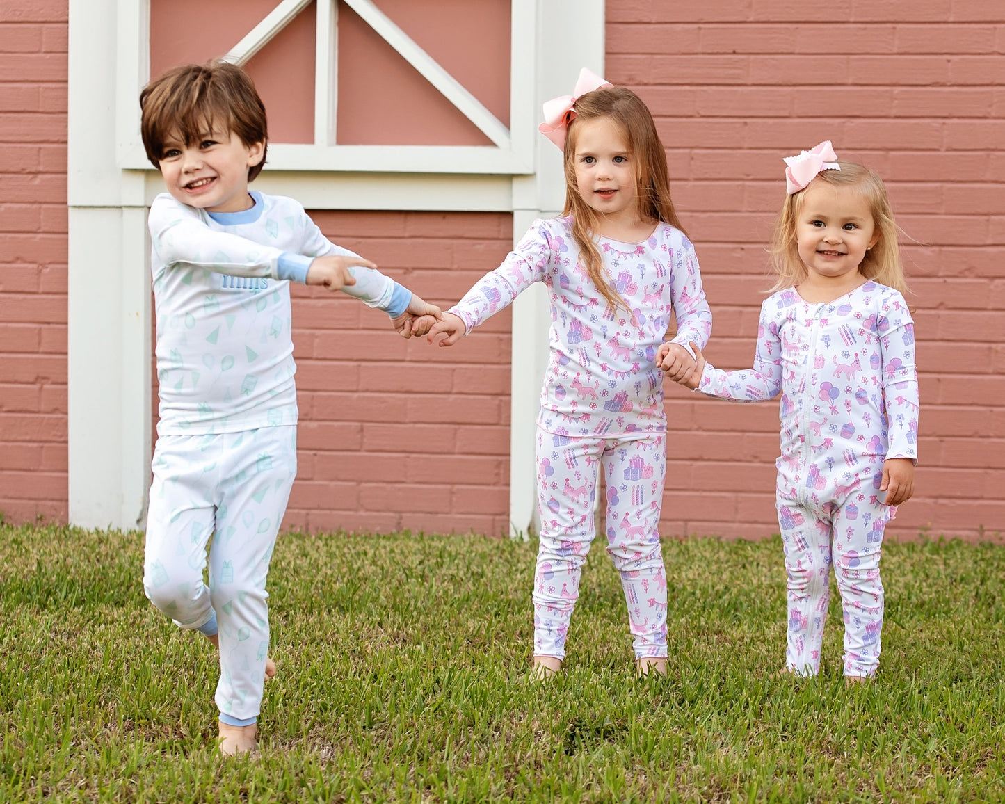 Three young children wearing matching birthday print pajamas — a boy in blue and green and two girls in pink and purple — holding hands and smiling while playing outside in front of a pink brick wall.