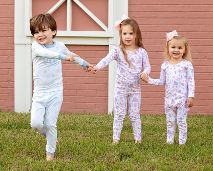 Three young children wearing matching birthday print pajamas — a boy in blue and green and two girls in pink and purple — holding hands and smiling while playing outside in front of a pink brick wall.