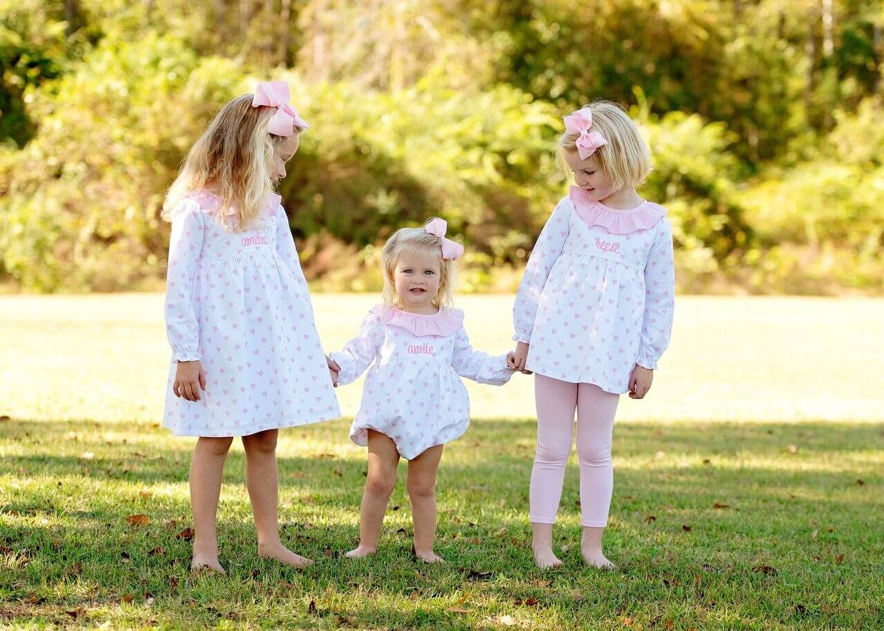 Three young girls in matching dresses standing outdoors on a grassy area with trees in the background.
