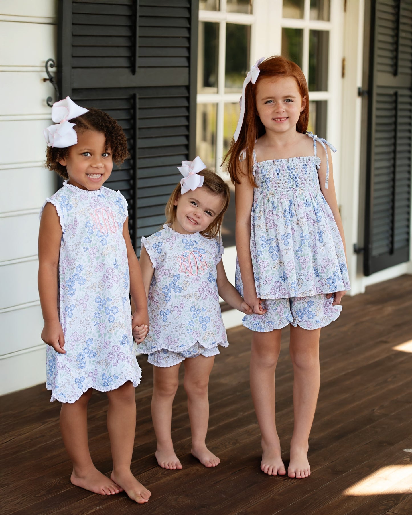 Three young girls in matching floral outfits, holding hands,  standing on a wooden deck.