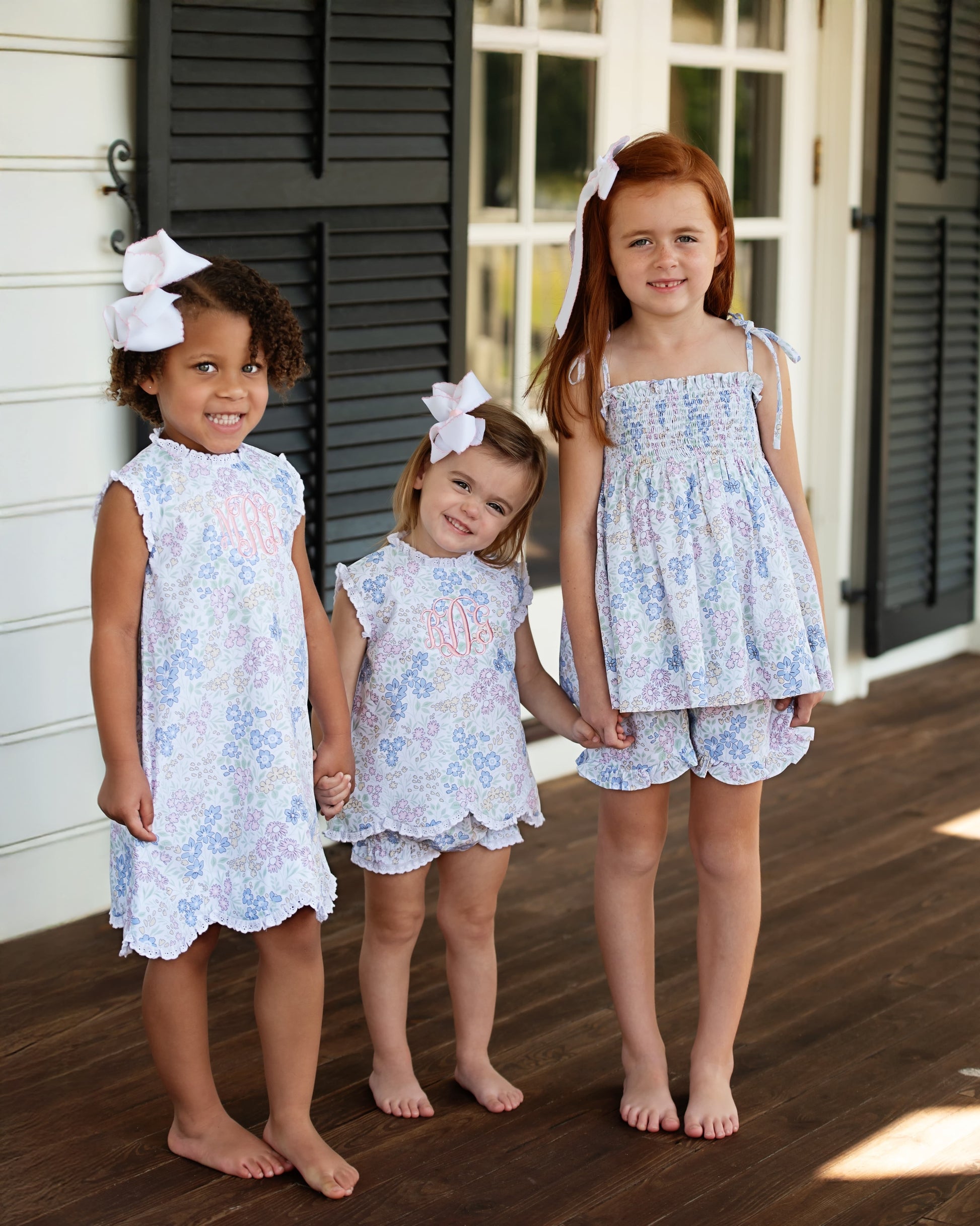Three young girls in matching floral outfits, holding hands while standing on a wooden deck.