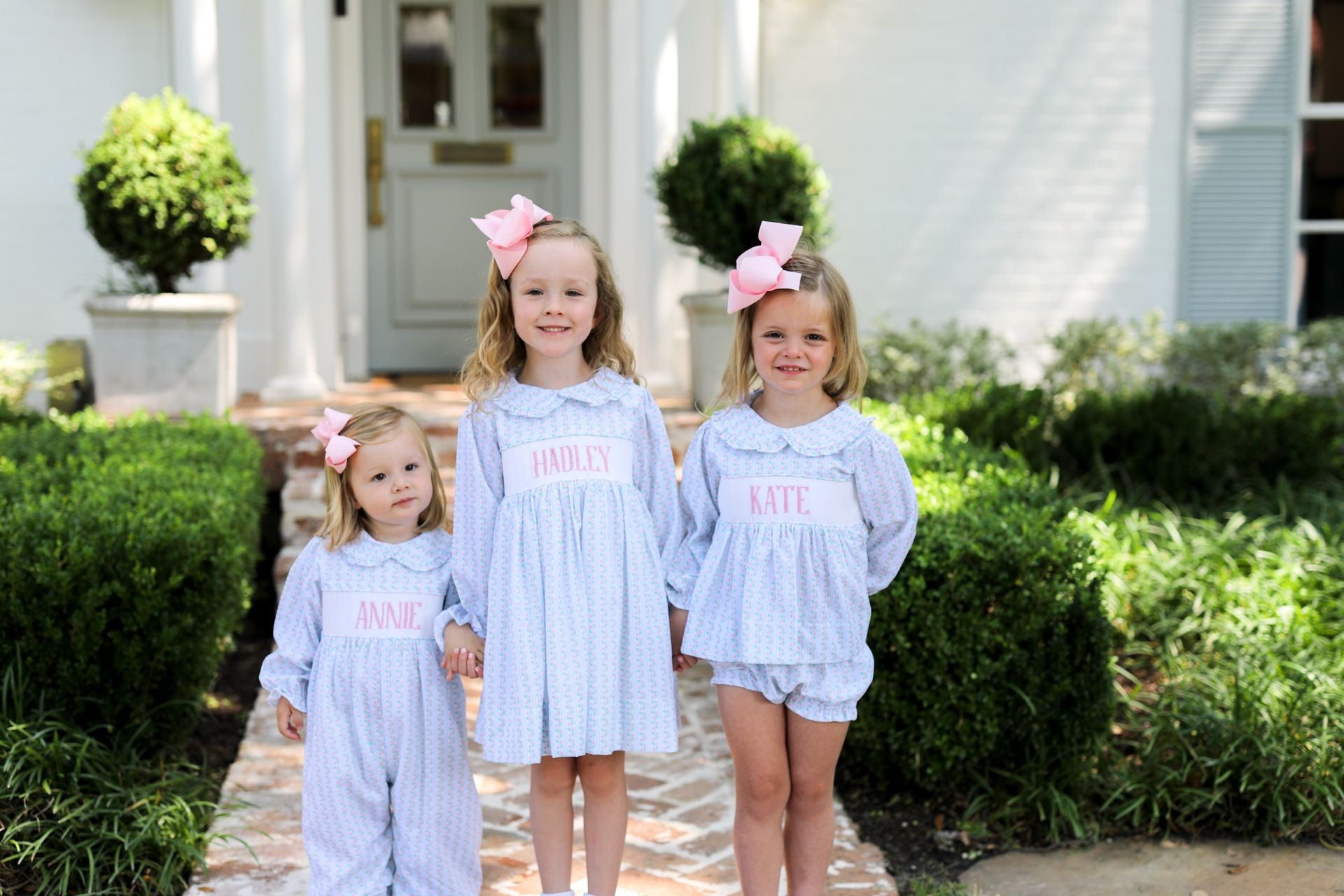 Three young girls in matching outfits with pink bows standing in front of a house.