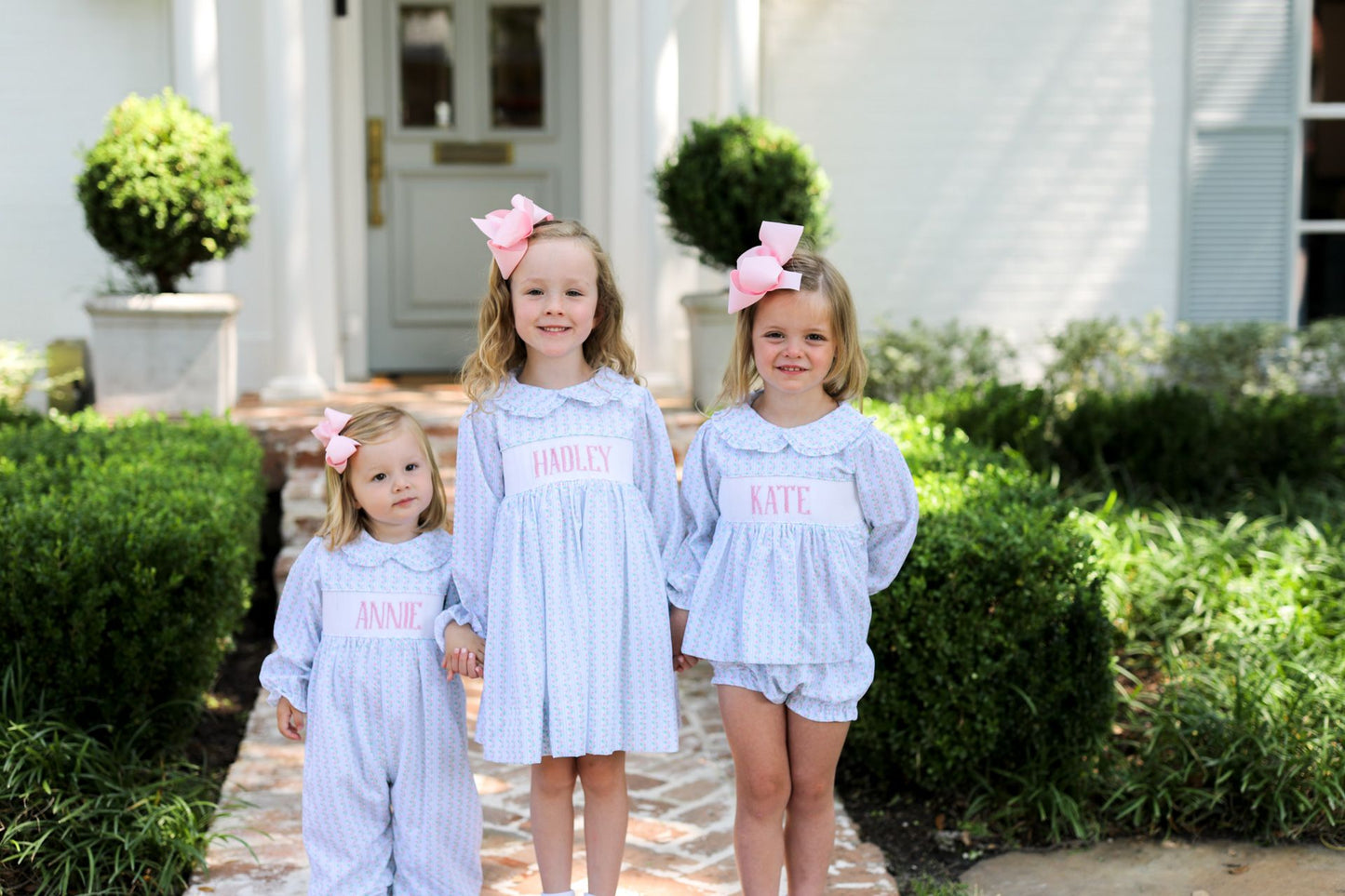 Three young girls in matching outfits with pink bows standing in front of a house.