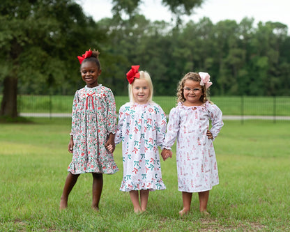 Three young girls standing on a grassy lawn outdoors, each wearing a long-sleeve dress with a different colorful print and a red or pink hair bow. Trees and a fence are visible in the background.