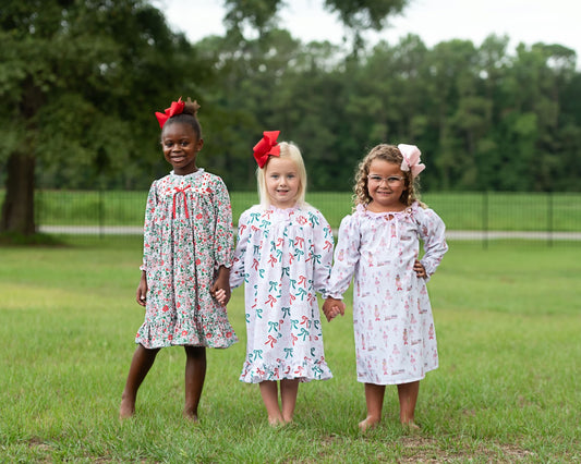 Three young girls standing on a grassy lawn outdoors, each wearing a long-sleeve dress with a different colorful print and a red or pink hair bow. Trees and a fence are visible in the background.