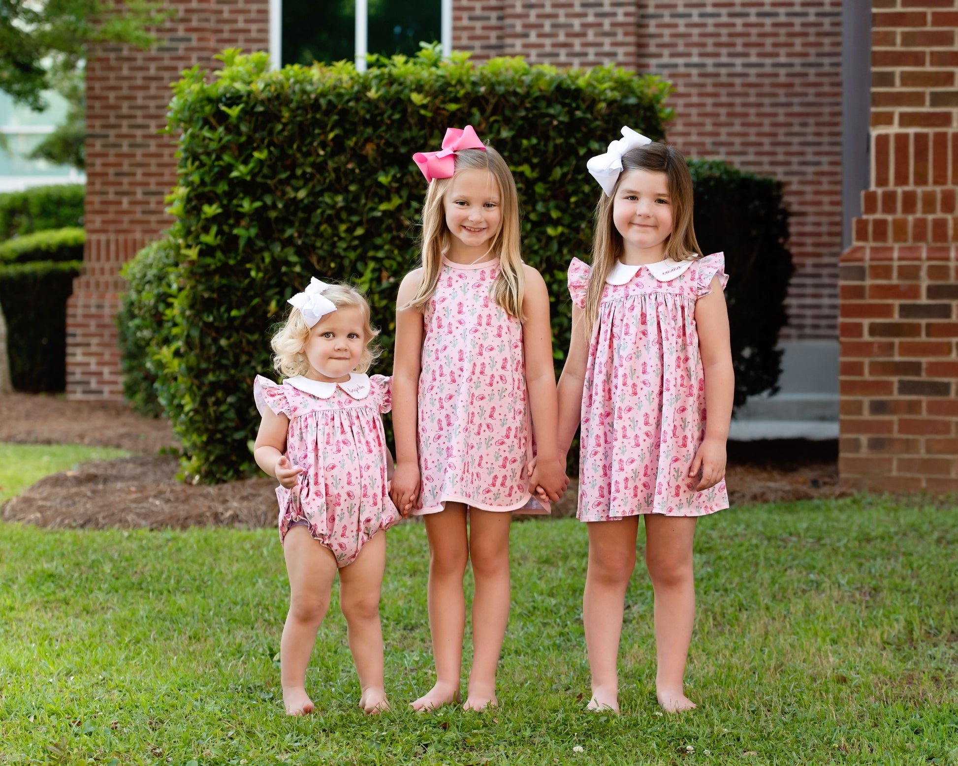 Three young girls wearing matching pink rodeo print dresses standing in front of a brick building.