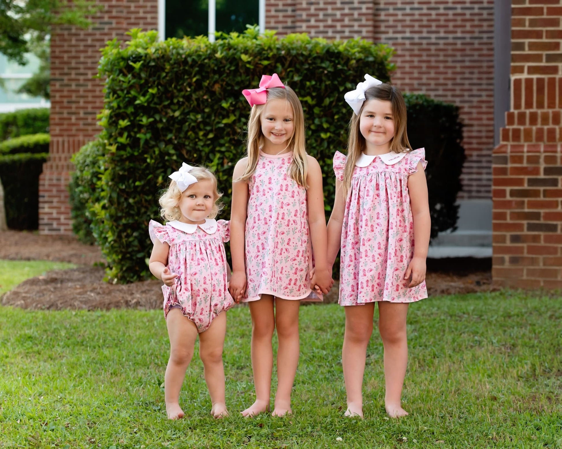 Three young girls wearing matching pink rodeo print dresses standing in front of a brick building.
