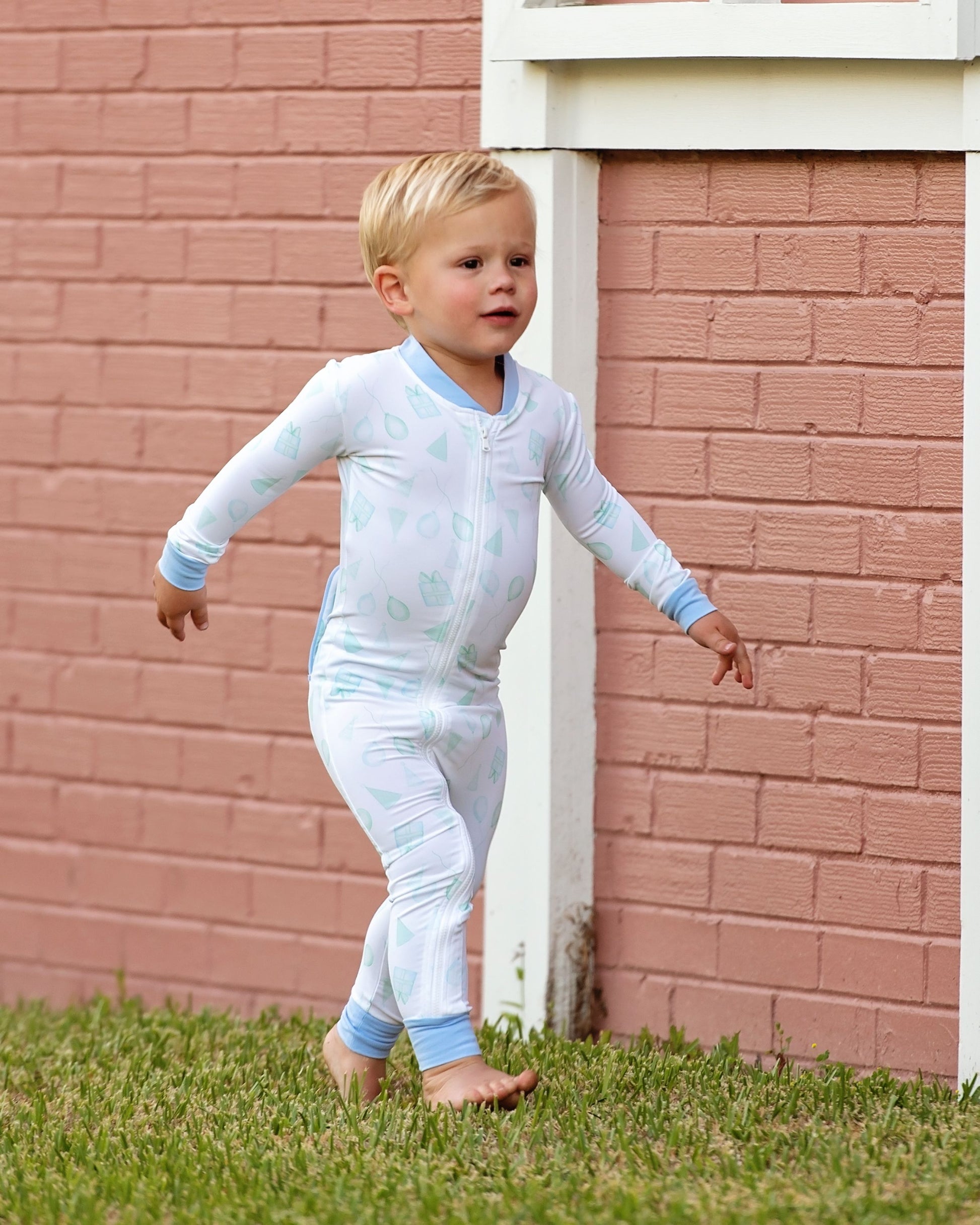 Toddler boy wearing blue and green birthday print zipper pajamas with light blue trim, walking barefoot on grass in front of a pink brick wall.