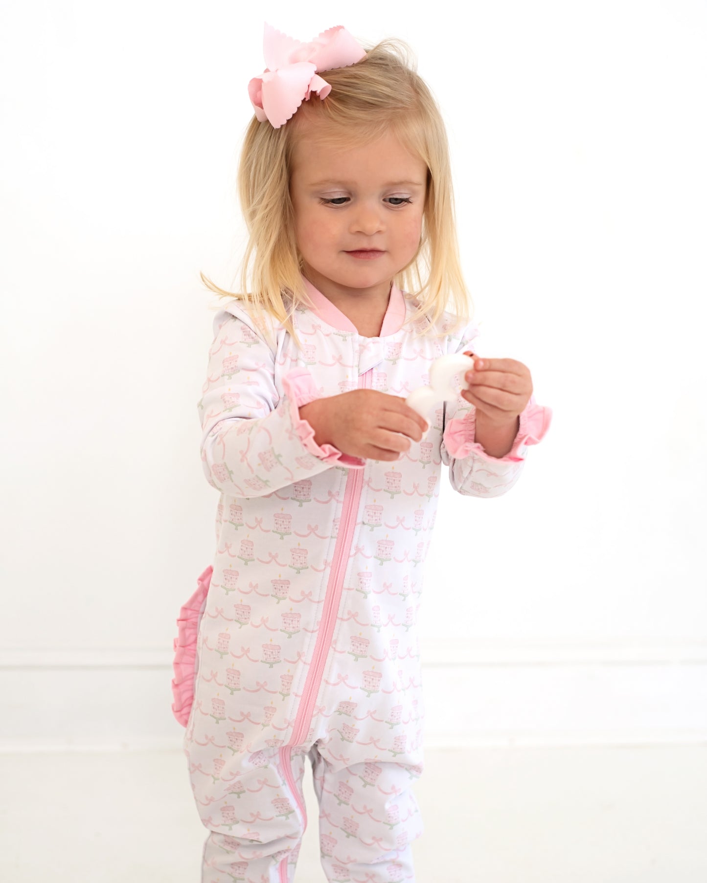 Toddler girl wearing pink and white birthday print zipper pajamas with ruffle details and a large pink bow, standing against a white background and holding a small toy.
