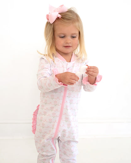Toddler girl wearing pink and white birthday print zipper pajamas with ruffle details and a large pink bow, standing against a white background and holding a small toy.