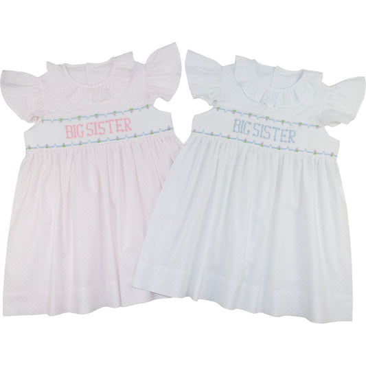 Two baby girl's smocked dresses with ruffled sleeves, shown side by side on a white background. The left dress is light pink with pink 'BIG SISTER' text and a small green/blue floral border.