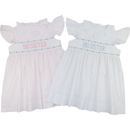 Two baby girl's smocked dresses with ruffled sleeves, shown side by side on a white background. The left dress is light pink with pink 'BIG SISTER' text and a small green/blue floral border.