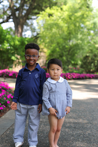 Two boys standing outdoors on a paved path in a garden. One boy wears a navy blue long-sleeve polo shirt with a small white monogram on the chest and matching blue-and-white checkered pants.