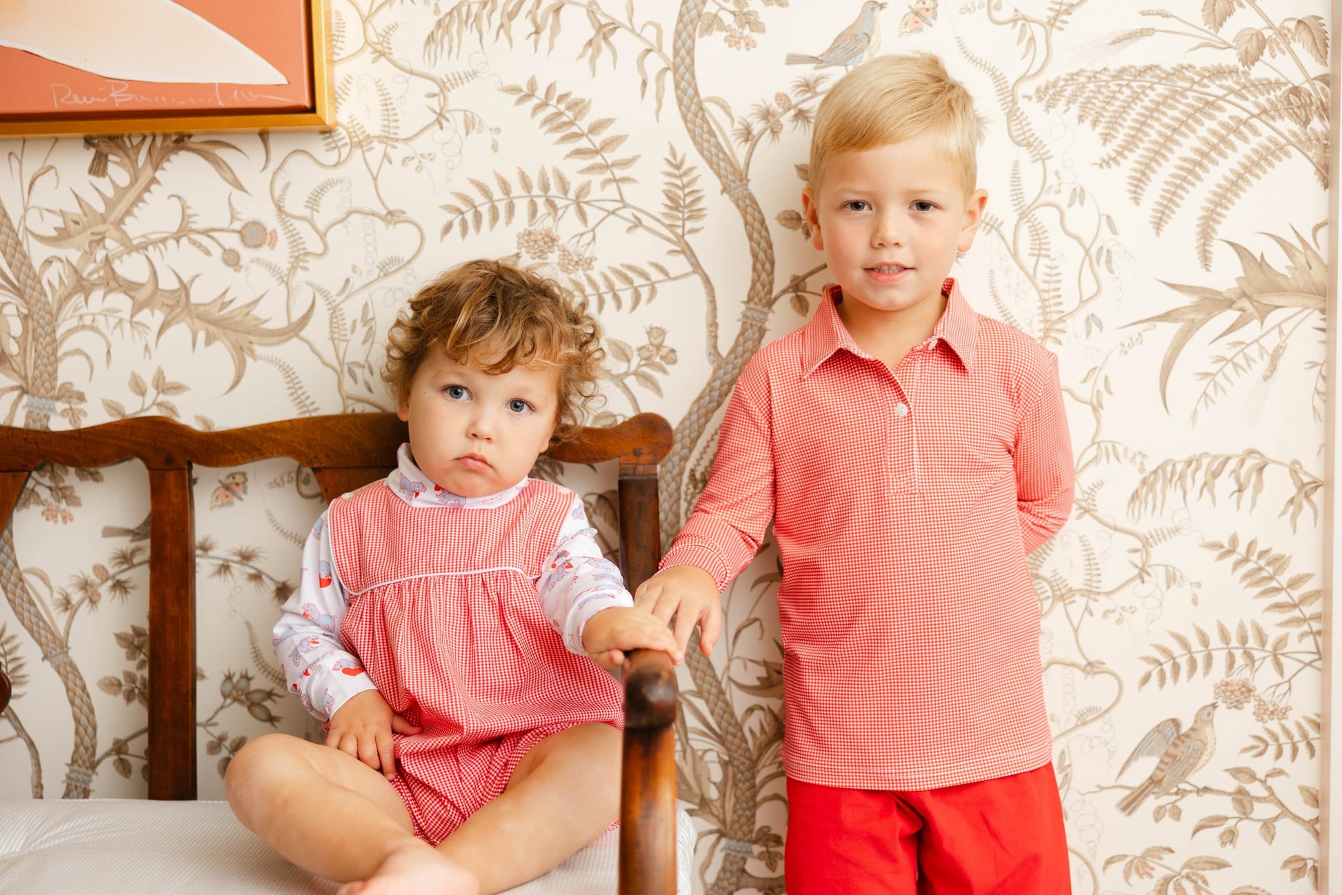 Two children, a boy and a girl, standing in front of a floral wallpapered wall.