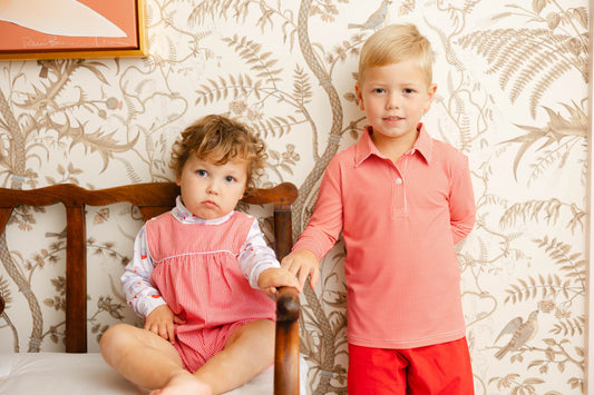 Two children, a boy and a girl, standing in front of a floral wallpapered wall.