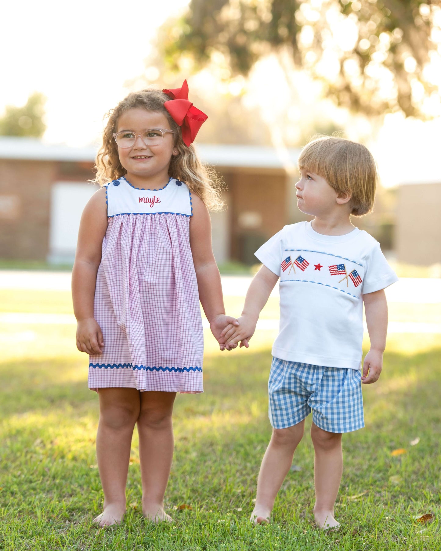 Two children holding hands outdoors, one in a dress with a red bow and the other in a white shirt with American flag design and blue shorts.