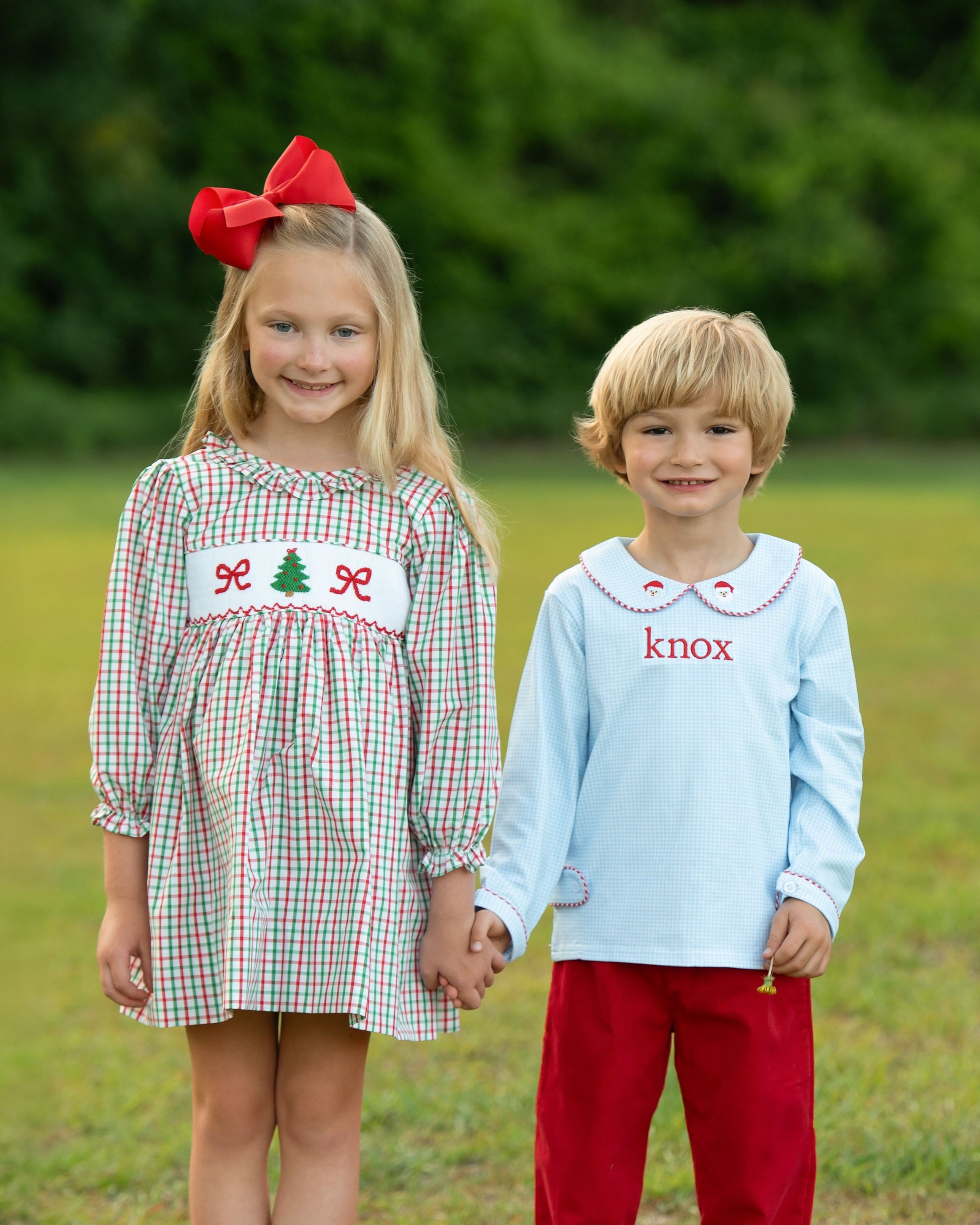 Two children holding hands outdoors, one in a dress with a tree design and the other in a light blue shirt with 'knox' printed on it.