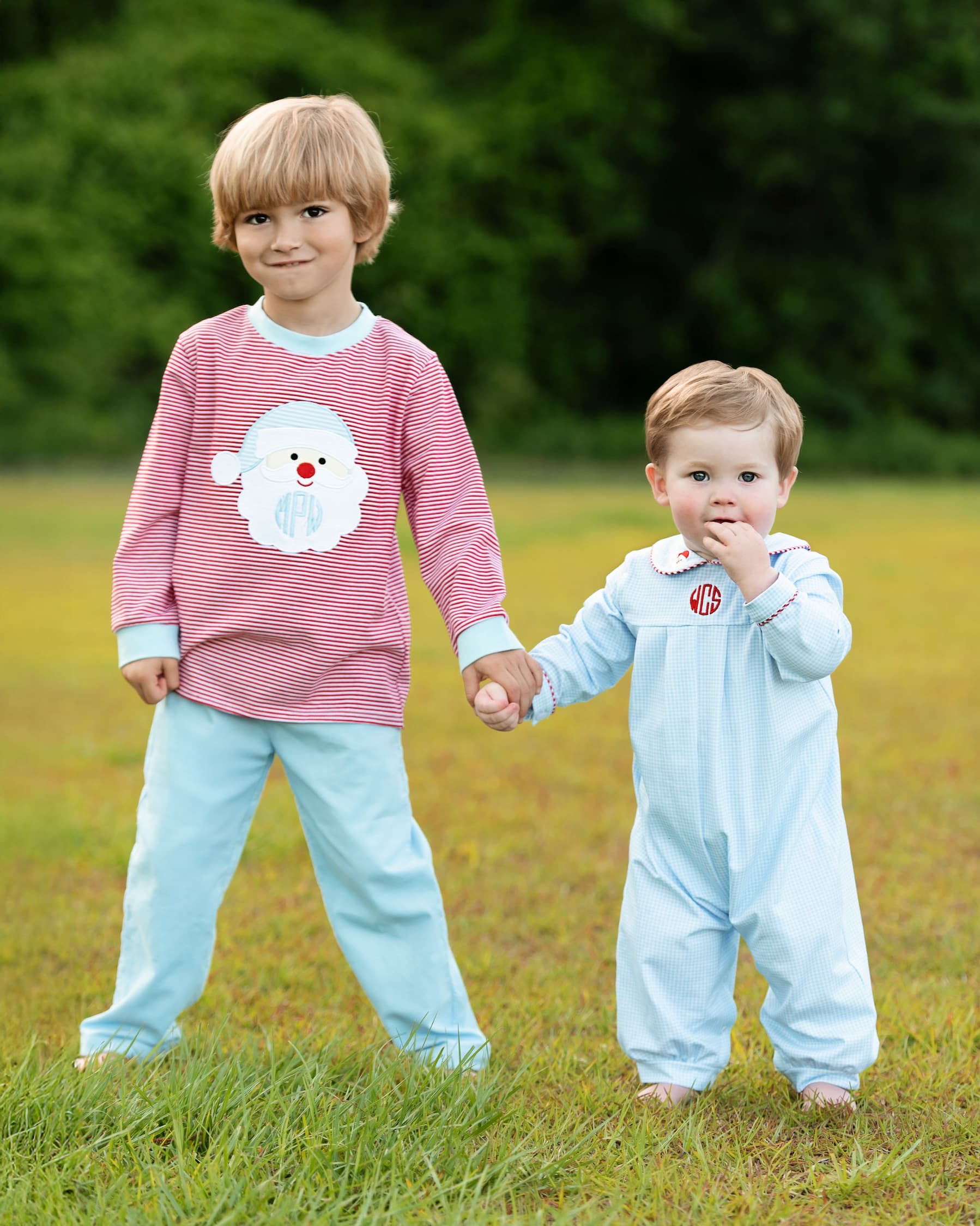 Two children holding hands outdoors, one wearing a pink sweater with a white dog design and the other in a light blue onesie.