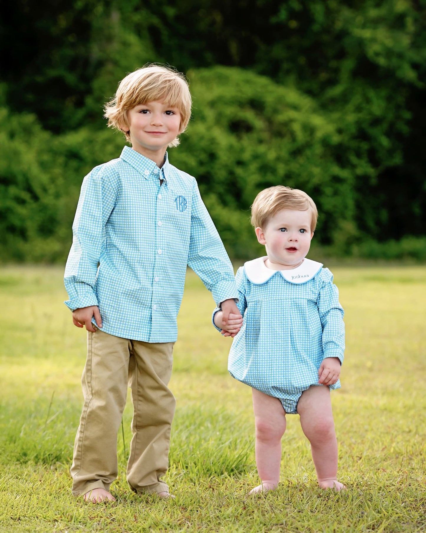 Two children in matching blue outfits standing in a grassy field with greenery in the background.