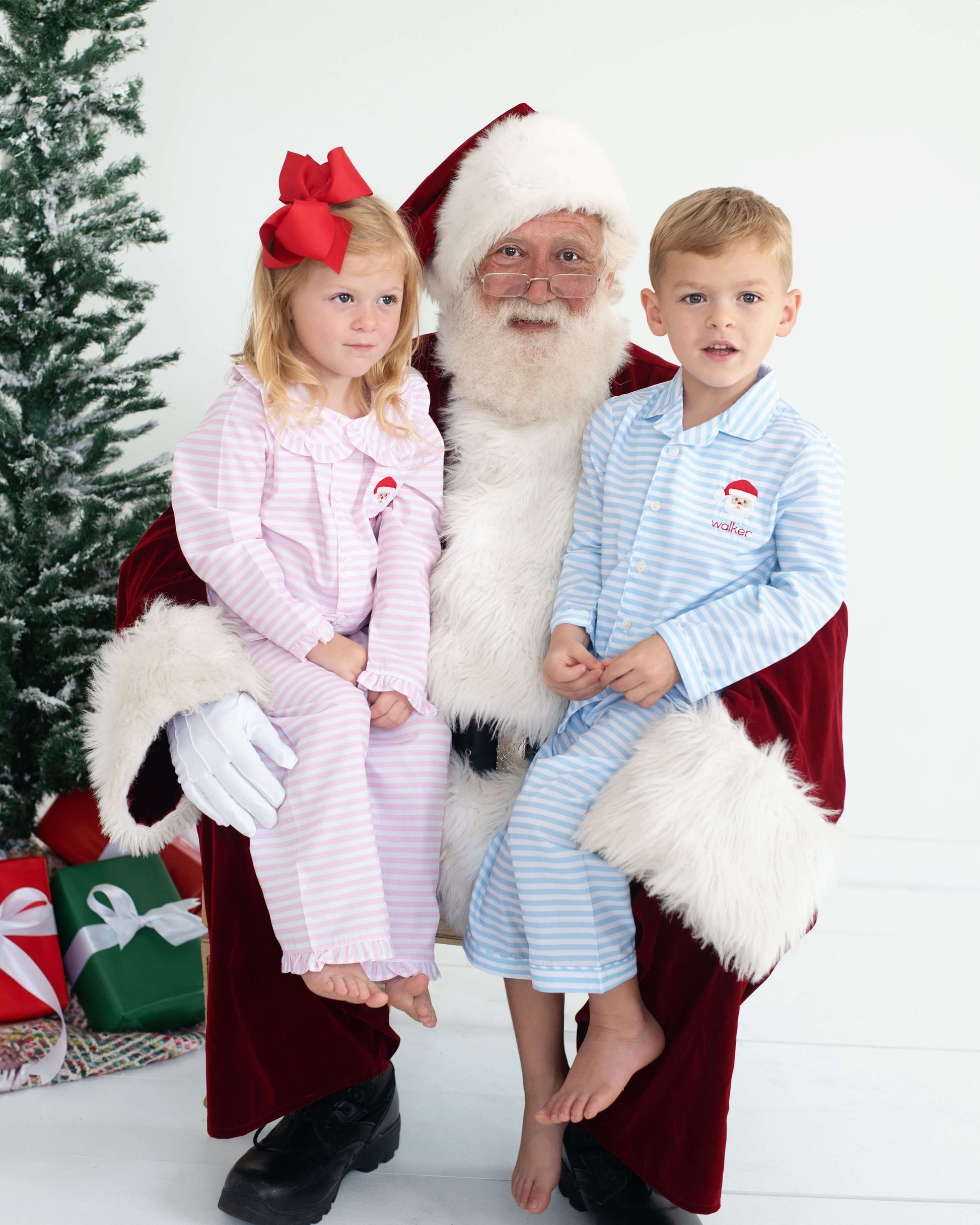 Two children in matching holiday pajamas sitting on Santa's lap with a Christmas tree and presents in the background.
