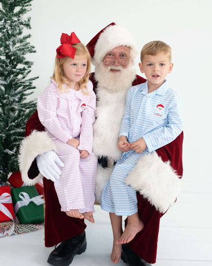 Two children in matching holiday pajamas sitting on Santa's lap with a Christmas tree and presents in the background.