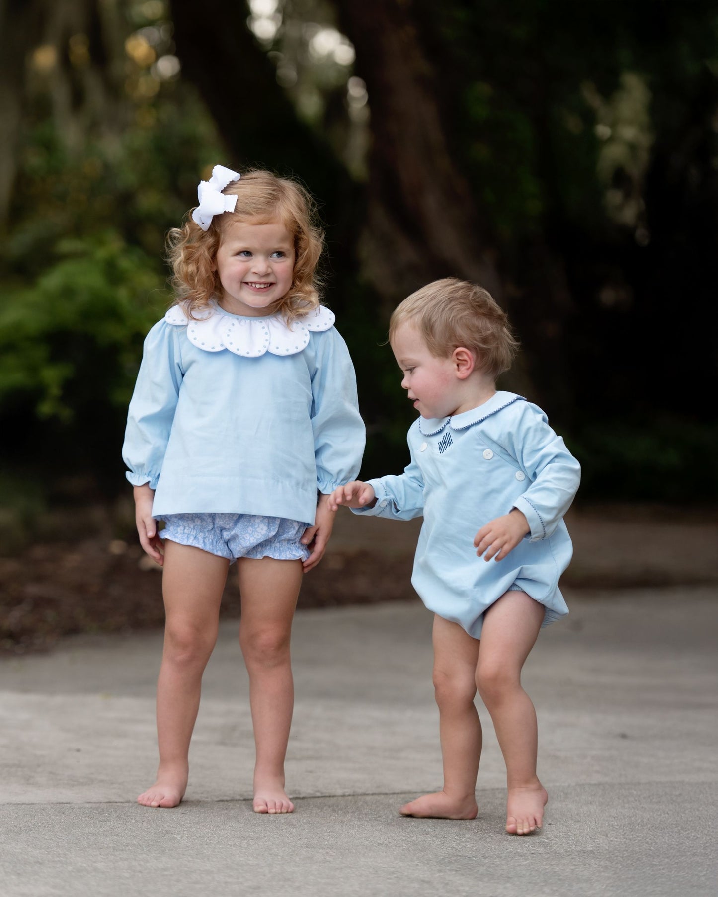 Two children in matching light blue outfits standing on a wooden path with greenery in the background.