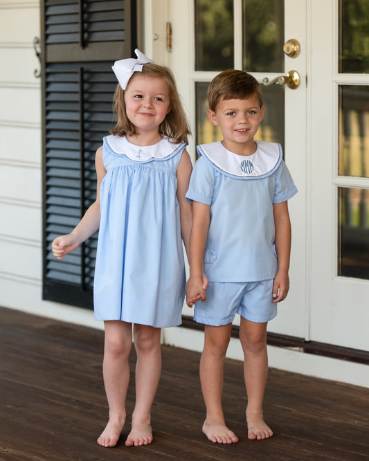 Two children in matching light blue outfits standing on a wooden porch holding hands.