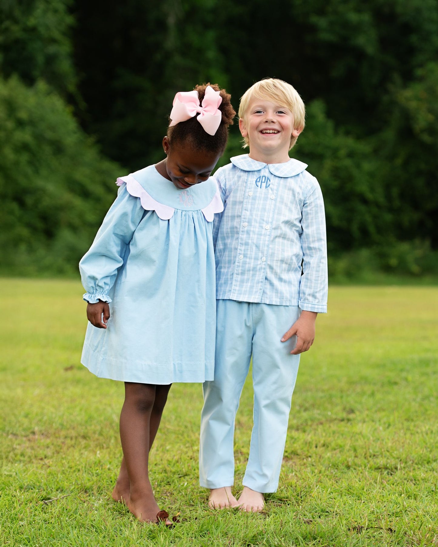 Two children in matching light blue outfits standing on grass with trees in the background