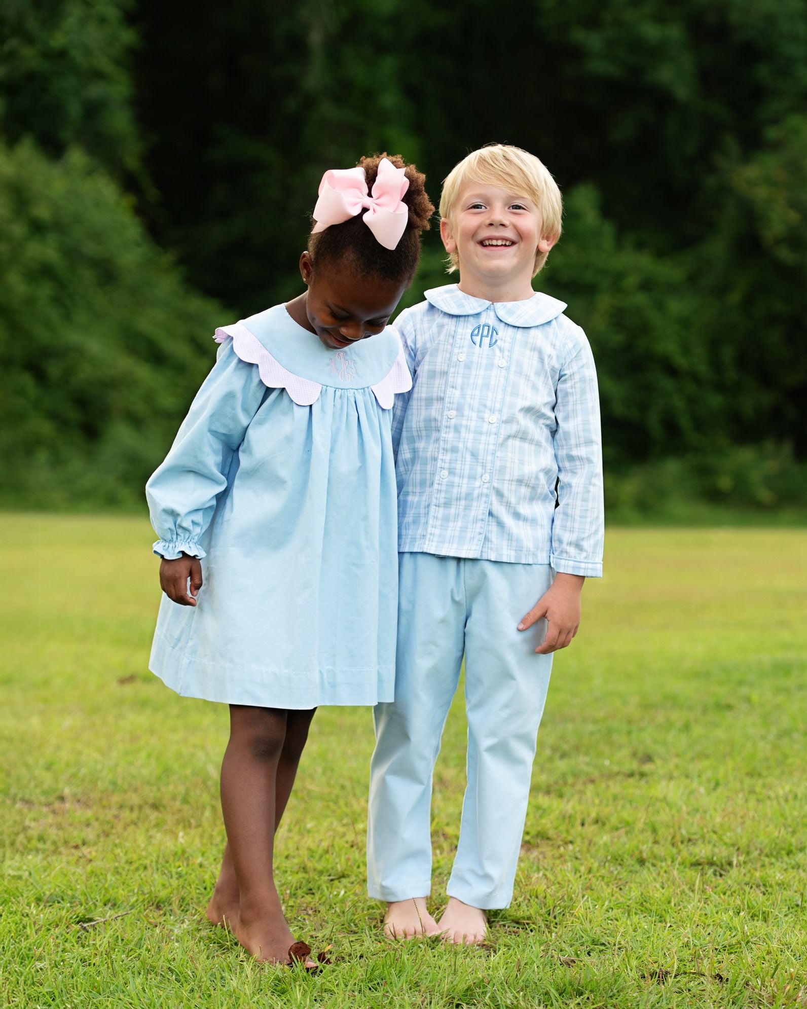 Two children in matching light blue outfits standing on grass with trees in the background