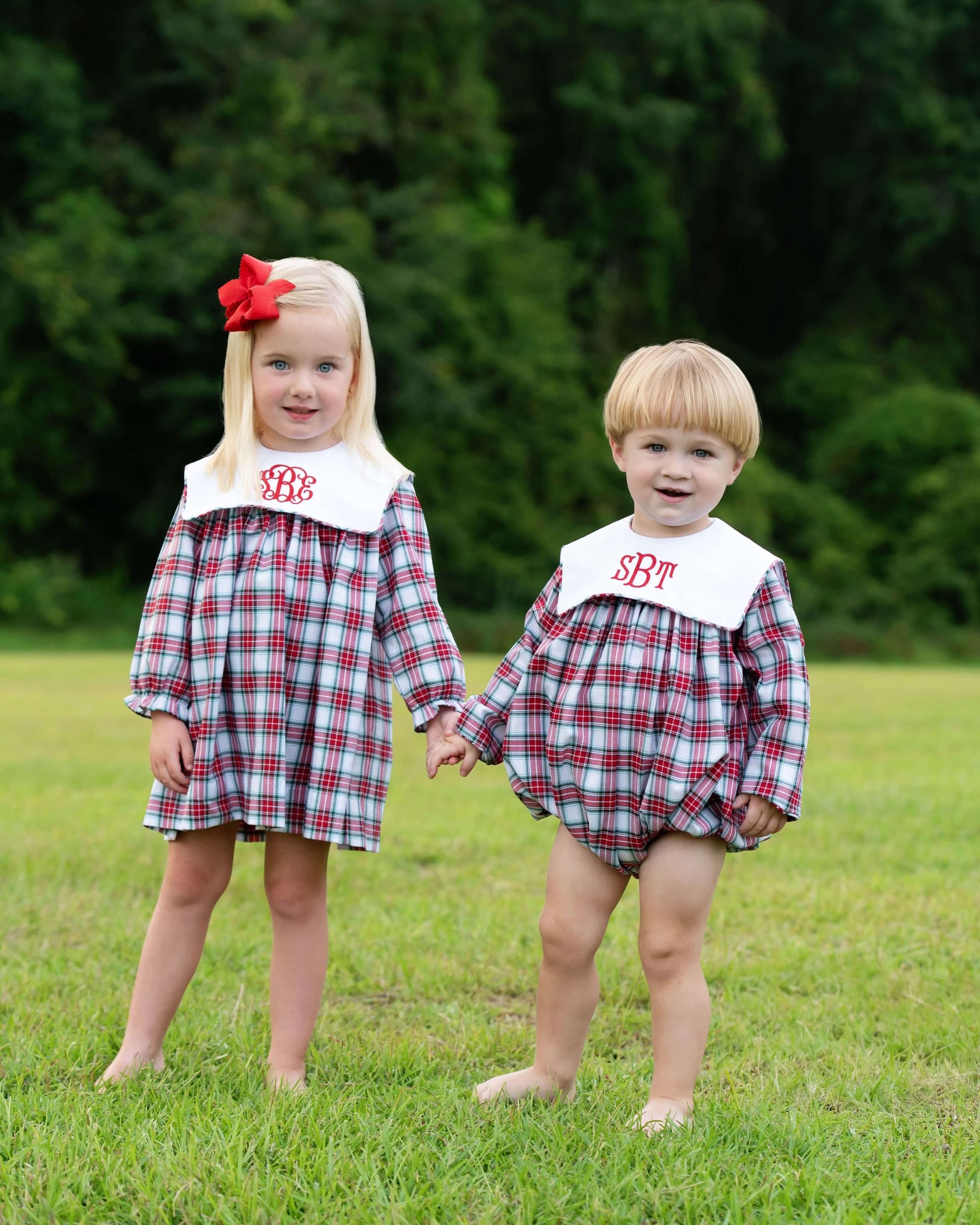 Two children in matching plaid outfits standing in a grassy area with trees in the background.