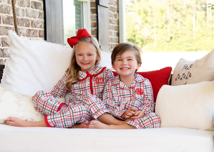 Two children in matching plaid pajamas sitting on a couch with pillows.