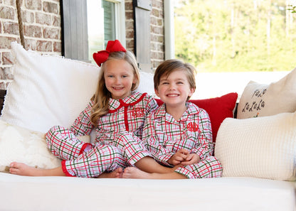 Two children in matching plaid pajamas sitting on a couch with pillows.